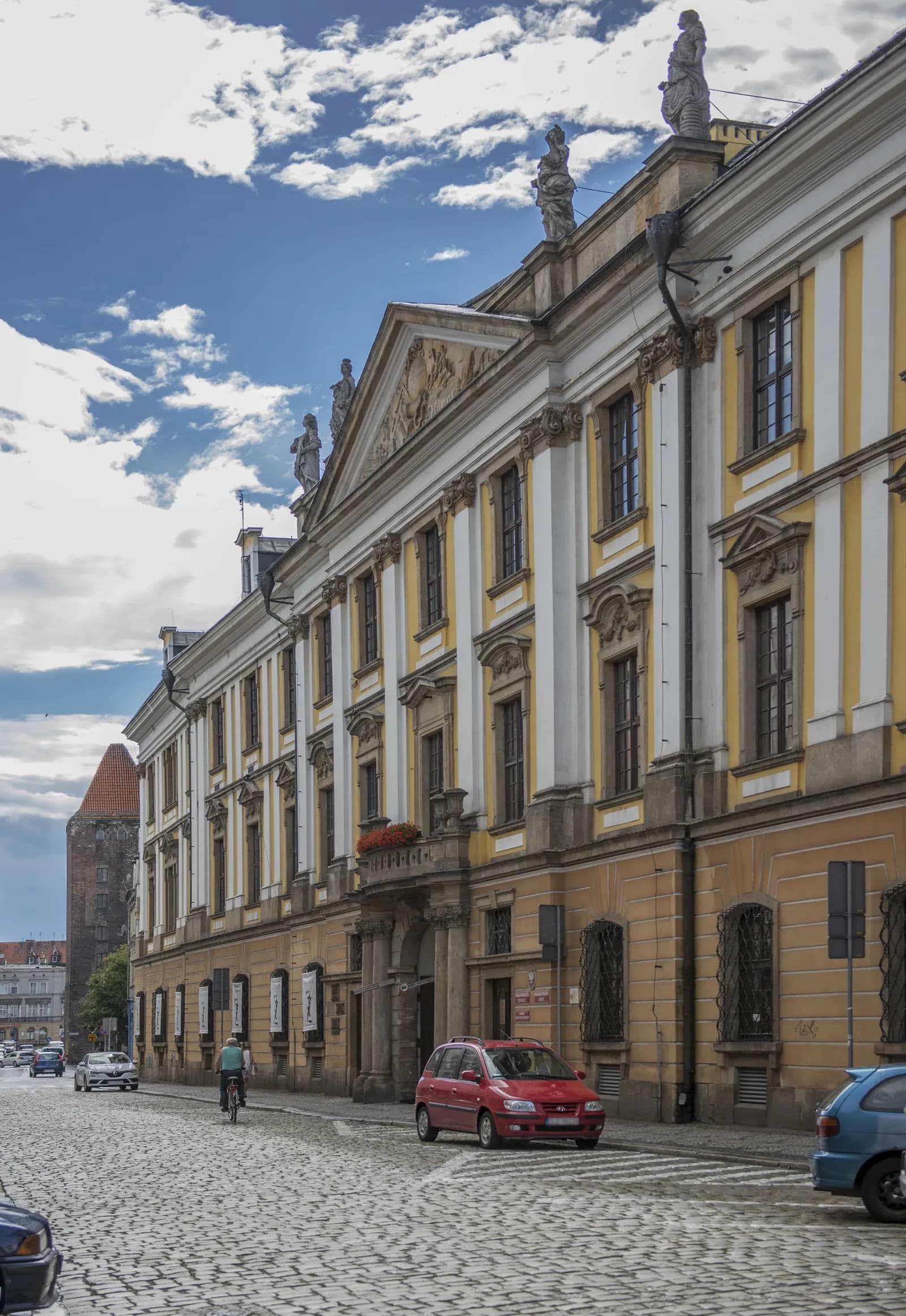 A grand, old yellow and white striped building with intricate architecture and statues on the roofline, with cars and a bicyclist on a cobblestone street in front.