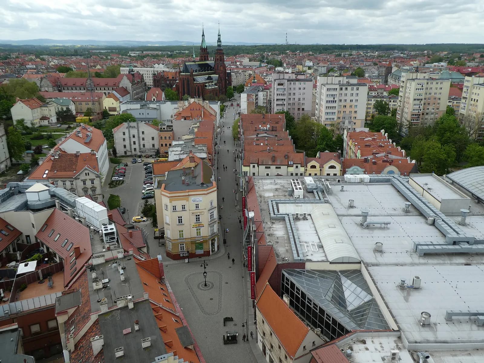 An aerial view shows a city street bordered by buildings with red-tiled roofs leading towards a large, dark church with two spires in the distance.